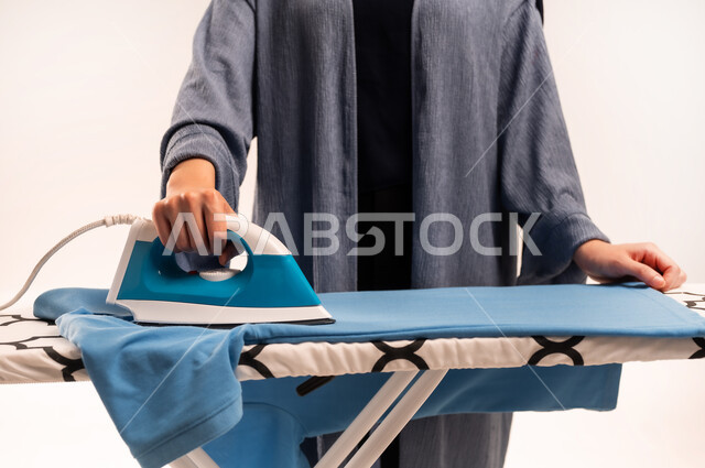 مفهوم التدبير المنزلي، تجفيف وتنظيم الملابس، استخدام الجهاز الكهربائي، انجاز المهام والأعمال الروتينية، بورتريه مقرب لايدي امرأة عربية خليجية سعودية محجبة ترتدي العباءة تقوم بكي الملابس باستخدام المكواة، خلفية بيضاء