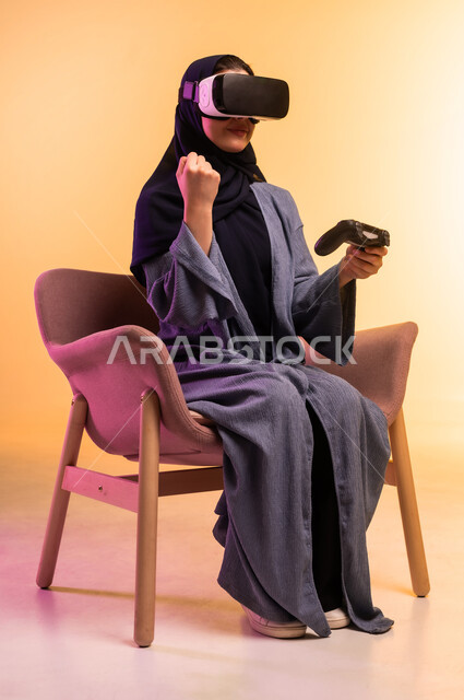 استخدام جهاز تقني حديث ومتطور، اللعب بالعاب الفيديو، بورتريه لامراة عربية خليجية سعودية محجبة ترتدي نظارة الواقع الافتراضي تجلس على كرسي مريح تحمل عصا التحكم ترفع يدها بتعابير النصر، ايماءات الاندماج والتركيز، التفاعل مع العالم الخيالي، خلفية ملونة