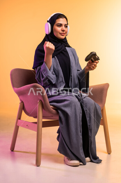 اللعب بألعاب الفيديو الترفيهية، الاستماع الى الموسيقى عبر الهيدفون، قضاء وقت ممتع في المنزل، بورتريه لإمرأة عربية خليجية سعودية محجبة تضع سماعة الرأس يجلس على كرسي مريح تحمل عصا التحكم ترفع يدها بايماءات لفوز والنصر، استخدام الجهاز التقني الحديث، خلفية ملونة