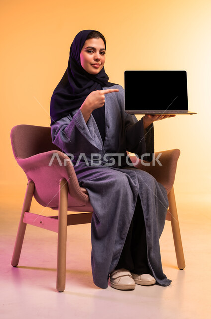 عرض شاشة سوداء فارغة عبر اللابتوب، ادارة العمل عن بعد، استخدام جهاز تقني حديث ومتطور, بورتريه لامرأة عربية خليجية سعودية محجبة ترتدي العباءة الرمادية تشير باصبع السبابة الى شاشة الحاسوب المحمول, إنجاز المهام المطلوبة عبر اللابتوب، خلفية ملونة