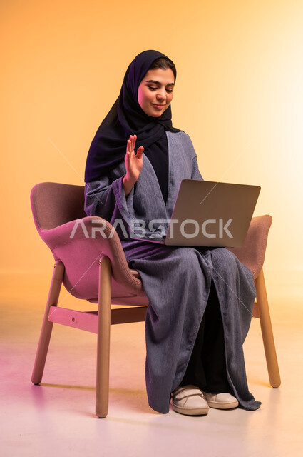 دمج التقنيات الحديثة والتكنولوجيا في الحياة العملية, انجاز المهام والأعمال, مفهوم العمل عن بعد,  بورتريه لامرأة عربية خليجية سعودية محجبة ترتدي العباءة الرمادية ترفع كف يدها بايماءات القاء التحية والسلام، استخدام جهاز الحاسوب المحمول, خلفية ملونة