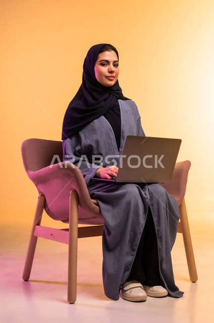 Accomplishing tasks and work, using a modern and advanced technical device, the concept of remote work, portrait of a veiled Saudi Arabian Gulf woman wearing a gray abaya working on a laptop, colorful background