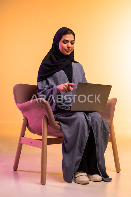 مفهوم العمل عن بعد, استخدام جهاز تقني حديث ومتطور، بورتريه لامرأة عربية خليجية سعودية محجبة ترتدي العباءة الرمادية تعمل على جهاز الحاسوب المحمول, انجاز المهام والأعمال, خلفية ملونة