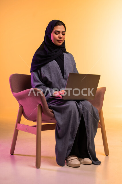 Using a modern and advanced technical device, the concept of remote work, a portrait of a veiled Saudi Arabian Gulf woman wearing a gray abaya working on a laptop, completing tasks and work, a colorful background