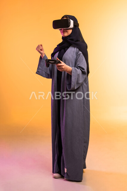 استخدام جهاز تقني حديث ومتطور، امراة عربية خليجية سعودية محجبة ترتدي نظارات ثلاثية الأبعاد تقف وتحمل بيدها عصا التحكم، التحول الرقمي والتقدم التقني، تعابير وايماءات الاندماج والتركيز في الواقع الافتراضي، الجسم بالطول الكامل، خلفية ملونة