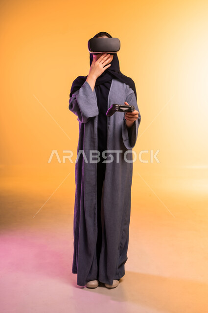 محاكاة العالم الخيالي، استخدام جهاز تقني حديث ومتطور، إمراة عربية خليجية سعودية محجبة ترتدي نظارات ثلاثية الأبعاد تقف وتحمل بيدها عصا التحكم، تعابير وايماءات الاندماج والتركيز في الواقع الافتراضي، الجسم بالطول الكامل، خلفية ملونة