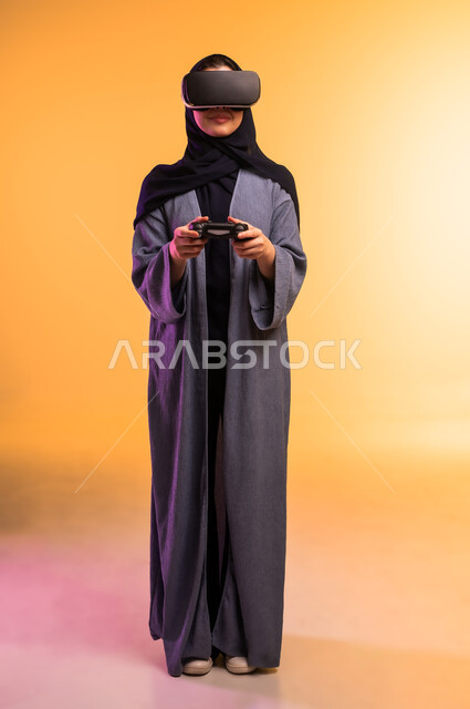 استخدام جهاز تقني حديث ومتطور، بورتريه لإمراة عربية خليجية سعودية محجبة ترتدي نظارات ثلاثية الأبعاد تقف وتحمل بيدها عصا التحكم، التحول الرقمي والتقدم التقني، تعابير وايماءات الاندماج والتركيز في الواقع الافتراضي، الجسم بالطول الكامل، خلفية ملونة