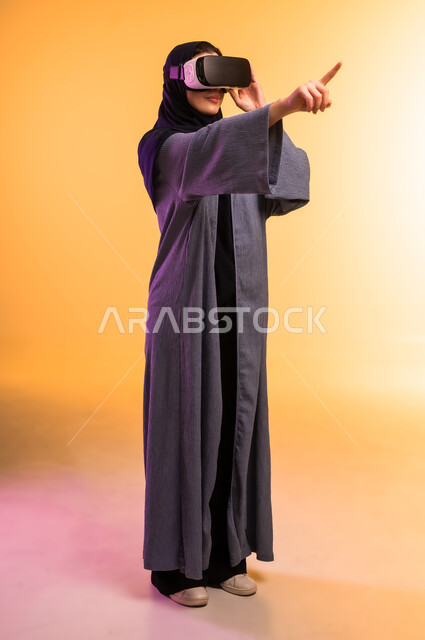 إيماءات التفاعل والاندماج مع العناصر الهولوجرامية، مفهوم التقنيات الإلكترونية الحديثة، بورتريه لإمراة عربية خليجية سعودية محجبة ترتدي العباءة ونظارات الواقع الإفتراضي (في آر) ثلاثية الأبعاد،  الجسم بالطول الكامل، خلفية ملونة