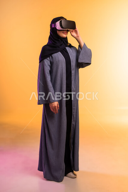 التحول الرقمي والتقدم التقني، إيماءات التفاعل مع العناصر الهولوجرامية، مفهوم التقنيات الإلكترونية الحديثة، بورتريه لإمراة عربية خليجية سعودية محجبة ترتدي العباءة ونظارات الواقع الإفتراضي (في آر) ثلاثية الأبعاد، الجسم بالطول الكامل، خلفية ملونة