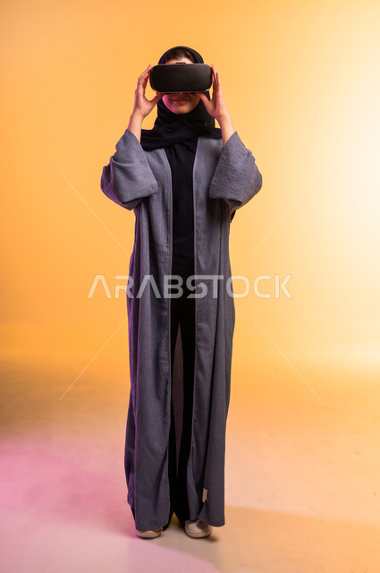 Concept of modern electronic technologies, gestures of interaction and integration with holographic elements, portrait of a veiled Saudi Arabian Gulf woman wearing an abaya and 3D virtual reality (VR) glasses, full-length body, colored background