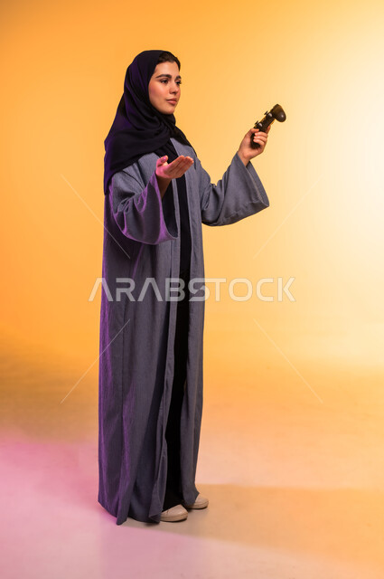 رفع كف اليد بايماءات الخسارة والاستفهام،  قضاء وقت ممتع في المنزل، اللعب بألعاب الفيديو الترفيهية، بورتريه لإمرأة عربية خليجية سعودية محجبة ترتدي العباءة تحمل بيدها عصا التحكم، الجسم بالطول الكامل، خلفية ملونة