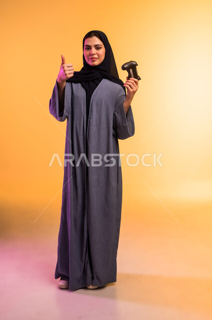 قضاء وقت ممتع في المنزل، لعب ألعاب الفيديو الترفيهية في عطلة نهاية الاسبوع،  بورتريه لإمرأة عربية خليجية سعودية محجبة ترتدي العباءة تحمل عصا التحكم وترفع اصبع الابهام بإيماءات الاعجاب، استخدام الجهاز التقني الحديث، الجسم بالطول الكامل، خلفية ملونة