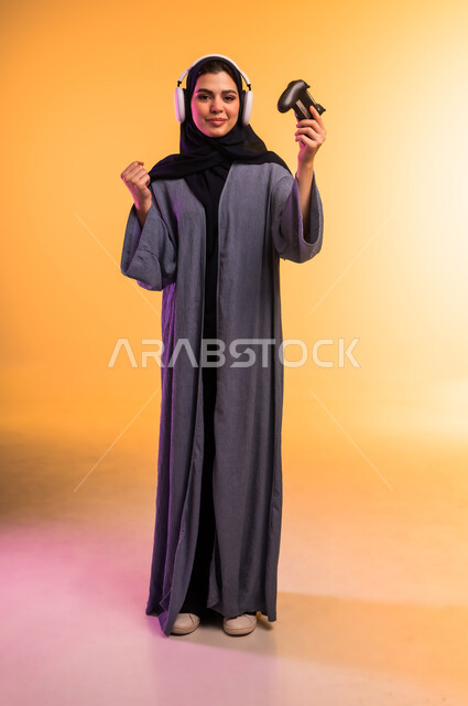 اللعب بألعاب الفيديو الترفيهية، قضاء وقت ممتع في المنزل، الاستماع الى الوسيقا عبر الهيدفون، بورتريه لإمرأة عربية خليجية سعودية محجبة تضع سماعة الرأس تحمل عصا التحكم ترفع يدها بايماءات لفوز والنصر، استخدام جهاز تقني حديث، الجسم بالطول الكامل، خلفية ملونة