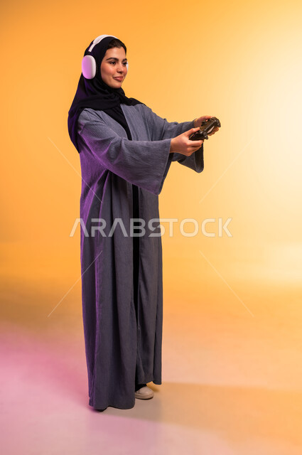 ايماءات التركيز والاندماج، الاستماع الى الوسيقا عبر الهيدفون، استخدام الجهاز التقني الحديث، قضاء وقت ممتع في المنزل، اللعب بألعاب الفيديو الترفيهية، بورتريه لإمرأة عربية خليجية سعودية محجبة تضع سماعة الرأس تحمل بيدها عصا التحكم، الجسم بالطول الكامل، خلفية ملونة