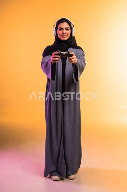 استخدام الجهاز التقني الحديث، الاستماع الى الوسيقا عبر الهيدفون، قضاء وقت ممتع في المنزل، اللعب بألعاب الفيديو الترفيهية، بورتريه لإمرأة عربية خليجية سعودية محجبة تضع سماعة الرأس تحمل بيدها عصا التحكم، ايماءات التركيز والاندماج، الجسم بالطول الكامل، خلفية ملونة