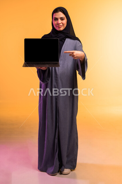 Blank black screen on laptop, remote work management, using modern and advanced technical device, portrait of a veiled Saudi Arabian Gulf woman wearing a gray abaya pointing with index finger at laptop screen, completing required tasks via laptop, colorful background