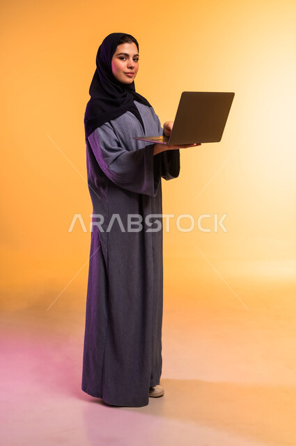 Remote work management, using modern and advanced technical device, portrait of a veiled Saudi Arabian Gulf woman wearing a gray abaya working on a laptop, completing required tasks via laptop, full body length, colored background
