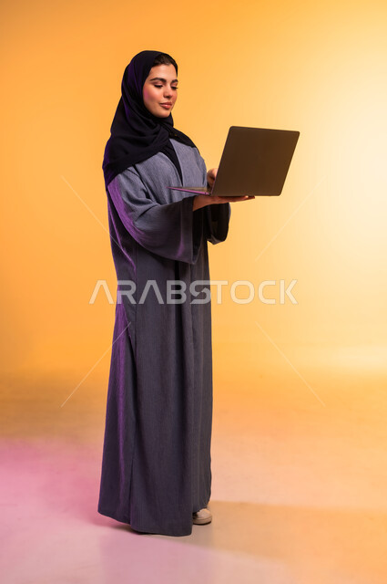 Completing required tasks via laptop, managing work remotely, using modern and advanced technical device, portrait of a veiled Saudi Arabian Gulf woman wearing a gray abaya working on a laptop, full body, colored background