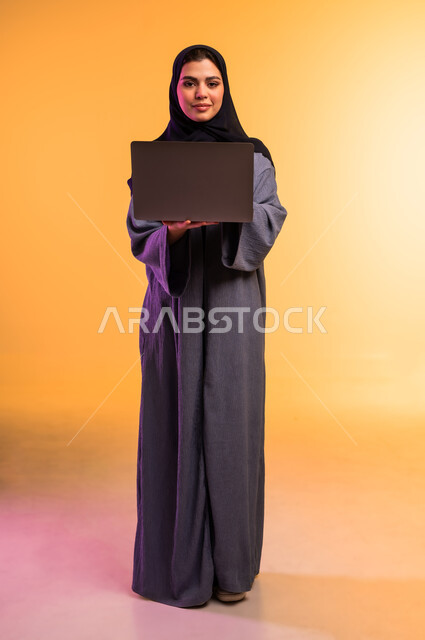 Using a modern and advanced technical device, managing work remotely, portrait of a veiled Saudi Arabian Gulf woman wearing a gray abaya working on a laptop, completing the required tasks via the laptop, full-length body, colored background