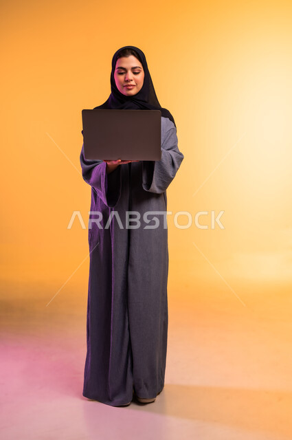 استخدام جهاز تقني حديث ومتطور, إنجاز المهام المطلوبة عبر اللابتوب، ادارة العمل عن بعد، بورتريه لامرأة عربية خليجية سعودية محجبة ترتدي العباءة الرمادية تعمل على جهاز الحاسوب المحمول, الجسم بالطول الكامل، خلفية ملونة