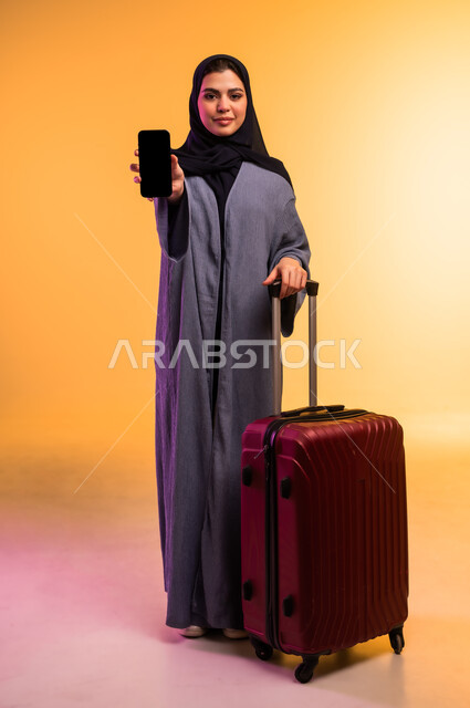 عرض شاشة سوداء فارغة عبر الهاتف المحمول، الذهاب في رحلة سفر، قضاء وقت ممتع بالاستماع الى الموسيقى, مفهوم الإصغاء والاستمتاع، استخدام الهيدفون في اوقات الفراغ، بورتريه لامرأة عربية خليجية سعودية ترتدي العباءة وتضع سماعات الرأس وبجانبها حقيبة السفر، خلفية ملونة