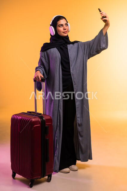 التقاط  صورة سيلفي عبر الهاتف المحمول، الذهاب في رحلة سفر، قضاء وقت ممتع بالاستماع الى الموسيقى, مفهوم الإصغاء والاستمتاع، استخدام الهيدفون في اوقات الفراغ، بورتريه لامرأة عربية خليجية سعودية ترتدي العباءة وتضع سماعات الرأس وبجانبها حقيبة السفر، خلفية ملونة