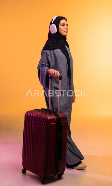 قضاء وقت ممتع بالاستماع الى الموسيقى, الاستعداد لقضاء الإجازة الصيفية والذهاب في رحلة سفر، مفهوم الإصغاء والاستمتاع، استخدام الهيدفون في اوقات الفراغ، بورتريه لامرأة عربية خليجية سعودية ترتدي العباءة وتضع سماعات الرأس وبجانبها حقيبة السفر، خلفية ملونة