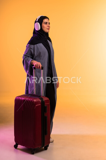 الاستعداد لقضاء الإجازة الصيفية والذهاب في رحلة سفر، قضاء وقت ممتع بالاستماع الى الموسيقى, مفهوم الإصغاء والاستمتاع، استخدام الهيدفون في اوقات الفراغ، بورتريه لامرأة عربية خليجية سعودية ترتدي العباءة وتضع سماعات الرأس وبجانبها حقيبة السفر، خلفية ملونة