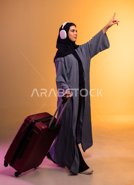 الذهاب في رحلة سفر، الاشارة باصبع السبابة إلى مكان ما، قضاء وقت ممتع بالاستماع الى الموسيقى, مفهوم الإصغاء والاستمتاع، استخدام الهيدفون في اوقات الفراغ، بورتريه من الجانب لامرأة عربية خليجية سعودية ترتدي العباءة وتضع سماعات الرأس وبجانبها حقيبة السفر، خلفية ملونة