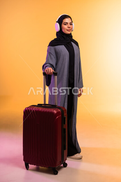 الاستعداد لقضاء الإجازة الصيفية والذهاب في رحلة سفر، قضاء وقت ممتع بالاستماع الى الموسيقى, مفهوم الإصغاء والاستمتاع، استخدام الهيدفون في اوقات الفراغ، بورتريه لامرأة عربية خليجية سعودية ترتدي العباءة وتضع سماعات الرأس وبجانبها حقيبة السفر، خلفية ملونة
