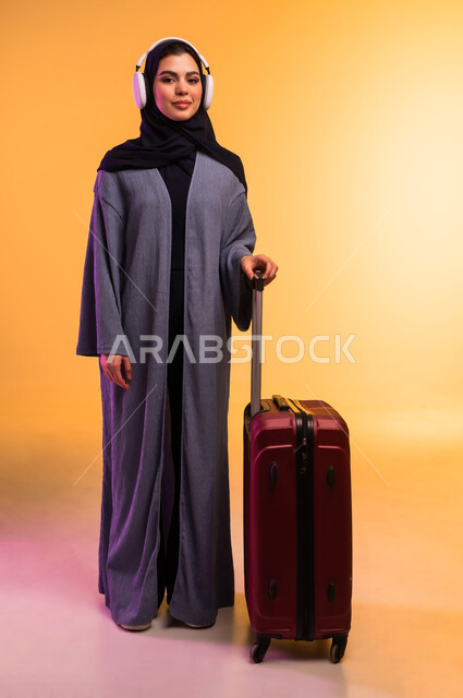 استخدام الهيدفون في اوقات الفراغ، الاستعداد لقضاء الإجازة الصيفية والذهاب في رحلة سفر، قضاء وقت ممتع بالاستماع الى الموسيقى, مفهوم الإصغاء والاستمتاع، بورتريه لامرأة عربية خليجية سعودية ترتدي العباءة وتضع سماعات الرأس وبجانبها حقيبة السفر، خلفية ملونة