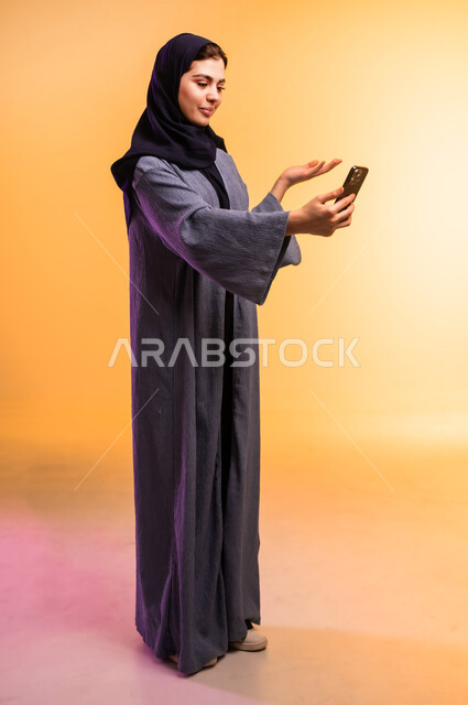 إجراء مكالمة فيديو مع الأهل والأصدقاء، بورتريه لشابة عربية خليجية سعودية ترتدي العباءة الملونة والحجاب تمسك بيدها الهاتف المحمول، استخدام جهاز تقني حديث ومتطور، رفع كف اليد للأعلى بايماءات السؤال وعدم المعرفة، الجسم بالطول الكامل، خلفية ملونة