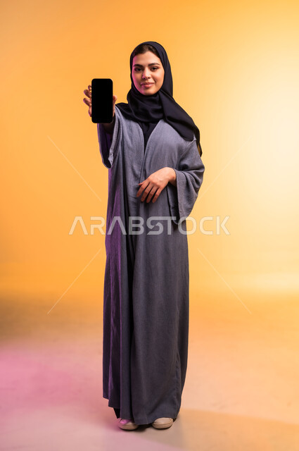Displaying a blank black screen via mobile, using a modern and advanced technical device, portrait of a veiled Saudi Arabian Gulf woman holding a mobile phone in her hand, looking at the camera with gestures of happiness and pleasure, full body, colored background