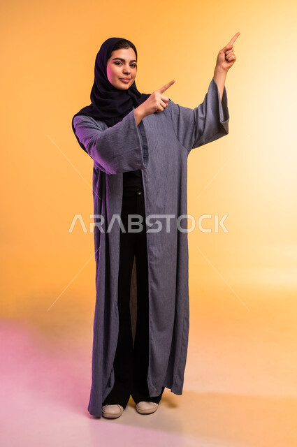 الإشارة إلى عنصر ما, تحديد موقع شيء معين, التوضيح والدلالة لمكان محدد, بورتريه لامرأة عربية خليجية سعودية محجبة ترتدي العباءة تشير بإصبعي السبابة، الجسم بالطول الكامل, خلفية ملونة