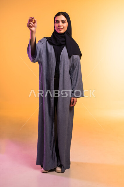 Taking notes, getting ready to write and mark something, portrait of a veiled Saudi Arabian Gulf woman wearing an abaya holding a pen in her hand writing something, concept of education with prescribed curricula, full length body, colored background