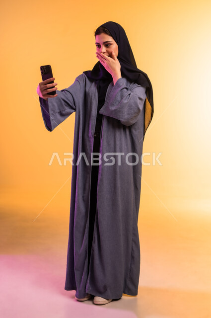 إيماءات التفاجؤ والذهول لسماع خبر ما، الاستغراب والانبهار والحماس, استخدام جهاز تقني حديث ومتطور، بورتريه لشابة عربية خليجية سعودية ترتدي العباءة والحجاب تضع يدها امام فمها وتمسك الهاتف المحمول, الجسم بالطول الكامل، خلفية ملونة