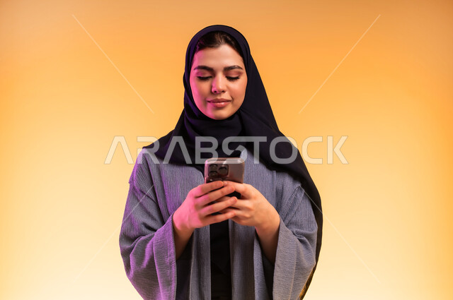 Gestures of focus and integration, chatting with family and friends, using a modern and advanced technical device, close-up portrait of a veiled Saudi Arabian Gulf woman holding a mobile phone, browsing social media via mobile, colorful background