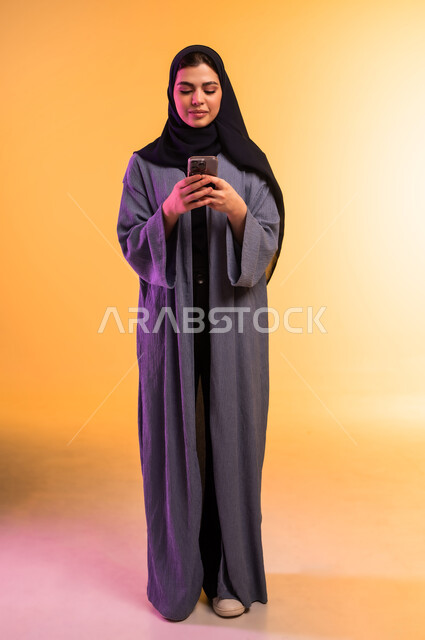 Using a modern and advanced technical device, gestures of focus and integration, chatting with family and friends, portrait of a veiled Saudi Arabian Gulf woman holding a mobile phone in her hand, browsing social media via mobile, full body, colorful background