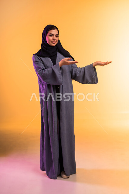 Lifting and carrying an item, caring for and taking care of one's appearance, portrait of a veiled Saudi Arabian Gulf woman wearing a colorful abaya raising her hands and pointing to a place, looking at the camera with gestures of happiness and pleasure, full body, colorful background