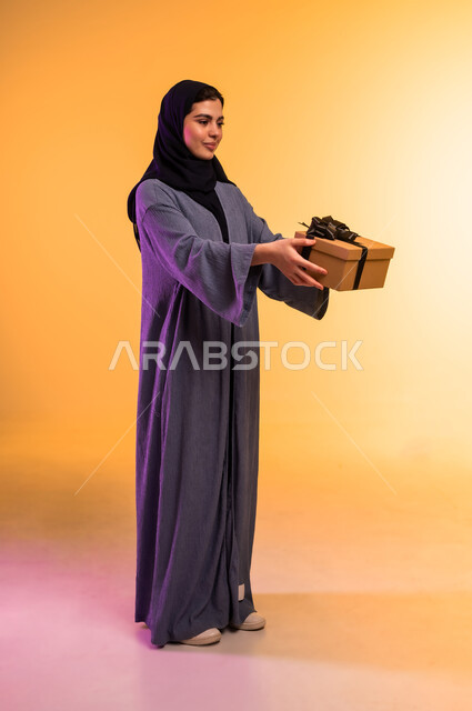 تقديم الهدايا في المناسبات السعيدة, الاحتفال بأعياد الميلاد, التعبير عن المحبة والتقدير, بورتريه لامرأة عربية خليجية سعودية محجبة ترتدي العباءة تمسك بيديها علبة هدية, النظر إلى الكاميرا بإيماءات السعادة والسرور, الجسم بالطول الكامل، خلفية ملونة