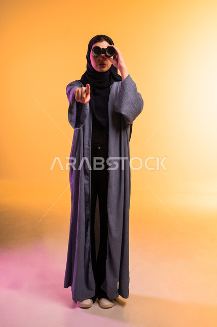الاشارة باصبع السبابة الى مكان ما، التخطيط المستقبلي للأهداف والطموحات، التفكير والتطلع إلى المستقبل، رؤية الأشياء البعيدة، حب الاستكشاف والمغامرة، بورتريه لشابة عربية خليجية سعودية محجبة ترتدي العباءة تمسك منظار ثنائي العينين، الجسم بالطول الكامل، خلفية ملونة
