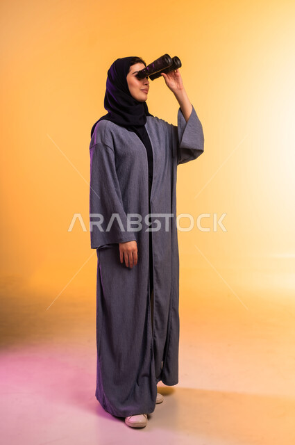 التخطيط المستقبلي للأهداف والطموحات، التفكير والتطلع إلى المستقبل، رؤية الأشياء البعيدة، حب الاستكشاف والمغامرة، بورتريه لشابة عربية خليجية سعودية ترتدي العباءة والحجاب تمسك منظار ثنائي العينين تنظر إلى شيء ما، الجسم بالطول الكامل، خلفية ملونة