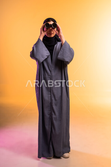 التفكير والتطلع إلى المستقبل، التخطيط المستقبلي للأهداف والطموحات، رؤية الأشياء البعيدة، حب الاستكشاف والمغامرة، بورتريه لشابة عربية خليجية سعودية ترتدي العباءة والحجاب تمسك منظار ثنائي العينين تنظر إلى شيء ما، الجسم بالطول الكامل، خلفية ملونة