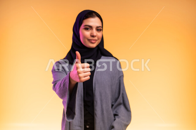 تعابير الرضا والموافقة, علامة الجودة والامتياز، مفهوم القبول والإعجاب,  بورتريه مقرب لامرأة عربية خليجية سعودية ترتدي العباءة تشير بإصبع الإبهام للأعلى، النظر إلى الكاميرا بإيماءات السعادة والسرور, خلفية ملونة