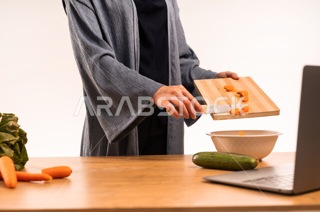 تعلم وصفات جديدة عبر الحاسوب المحمول، استخدام لوح التقطيع الخشبي، بورتريه مقرب لامراة عربية خليجية سعودية ترتدي العباءة تقف خلف طاولة عليها مجموعة من الخضراوات الطازجة في المطبخ، اتباع نظام غذائي صحي، إعداد السلطات والأطباق المفيدة، خلفية بيضاء