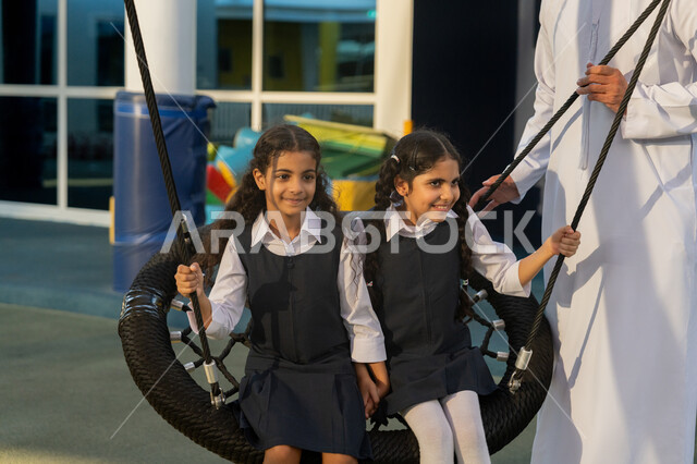 نشاط ترفيهي مسلي في مدارس الإمارات العربية المتحدة، الشعور بالفرح والسرور، قضاء وقت ممتع في اللعب واللهو مع الأصدقاء، فتاتان عربيتان خليجيتان اماراتيتان ترتديان الزي المدرسي تجلسان على الأرجوحة في ملعب المدرسة يقف بجانبهما المعلم
