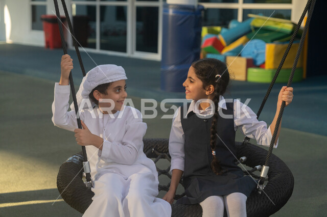 نشاط ترفيهي في مدرسة في الإمارات العربية المتحدة، قضاء وقت ممتع في اللعب واللهو مع الأصدقاء، ولد عربي خليجي اماراتي يرتدي الكندورة والعمامة يجلس على الارجوحة مع زميلته في الباحة المدرسية، الشعور بالفرح والسرور