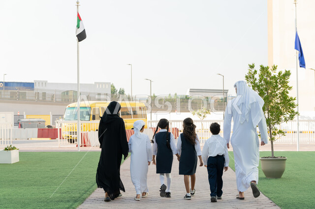 إعطاء الدعم النفسي والتحفيز المعنوي للطلاب، انتهاء الدوام المدرسي، موسم العودة إلى المدارس في الامارات العربية المتحدة، صورة من الخلف لمعلم ومعلمة عربيان خليجيان اماراتيان يخرجان من المدرسة مع الطلاب، الحافلة المدرسية الصفراء