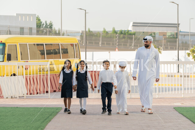 الاستعداد لبدء العام الدراسي الجديد بتعابير السرور، إعطاء الدعم النفسي والتحفيز المعنوي للطلاب، موسم العودة إلى المدارس في الامارات العربية المتحدة، معلم عربي خليجي اماراتي يرتدي الكندورة و الغترة يدخل الى المدرسة مع الطلاب، الحافلة المدرسية الصفراء