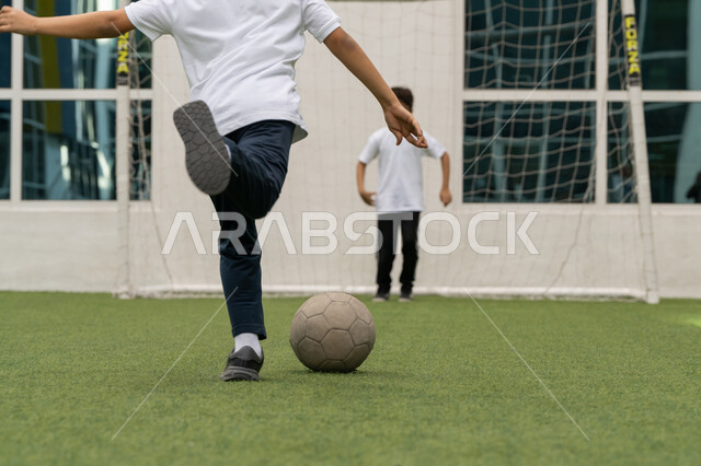اللعب في كرة القدم في ملعب المدرسة بإيماءات الحماس ,الشغف في ممارسة الرياضات المفضلة مع الأصدقاء, تعزيز القيم الاجتماعية والثقافية بين الطلاب, الاهتمام في الرياضة في الامارات العربية المتحدة, صورة من الخلف لطالب عربي خليجي اماراتي يقوم بتسديد الهدف بدقة
